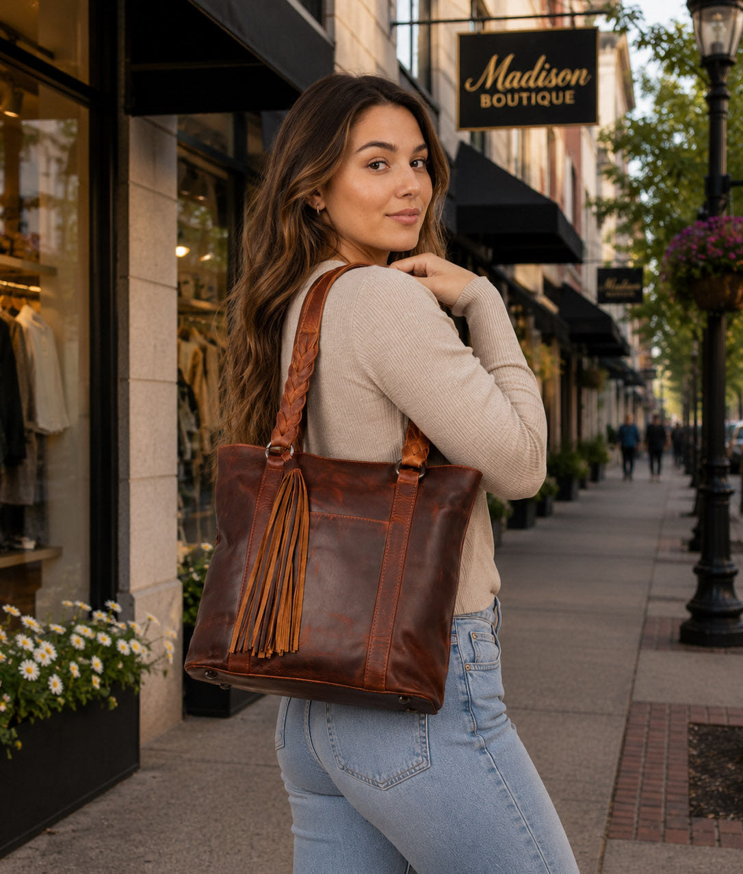 A woman stands on a city sidewalk holding then Concealed Carry Bella Leather Tote - Women's Purse for Pistol - Locking Purse with pistol slot - Leather Conceal Carry purse for Women - Concealed carry purse holster - Conceal Carry Handbag for Pistol - Handgun Bags for Womenwith long tassel straps. She is wearing a beige V-neck sweater and blue jeans.