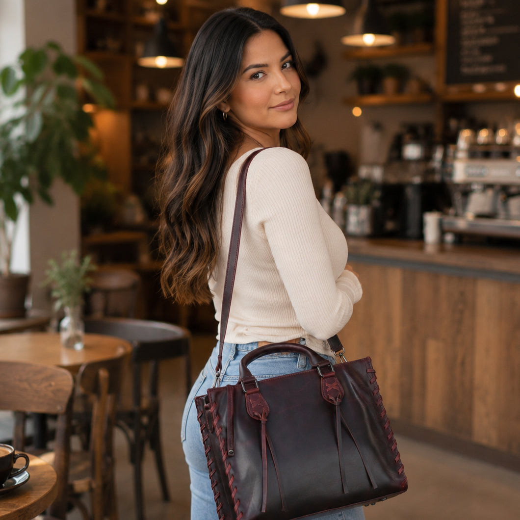 A woman in a cozy cafe setting wearing a cream ribbed long-sleeve top and light blue jeans, carrying a dark brown leather tote bag with burgundy stitching and top handles.