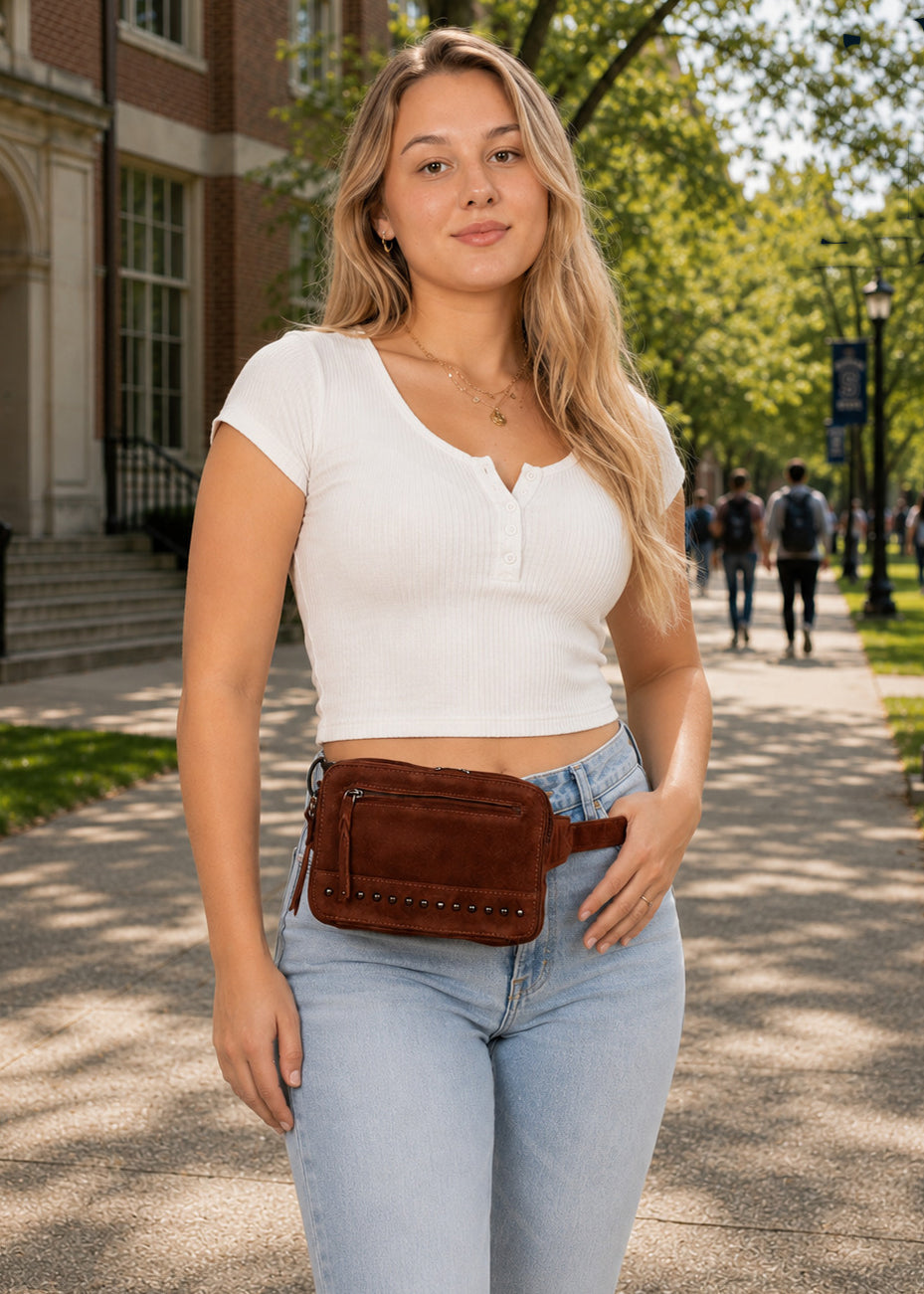 A woman wearing a brown leather fanny pack/belt bag with silver studs, styled with a white ribbed crop top and light blue jeans, standing on a sunny outdoor campus walkway. Concealed Carry Kailey Leather Purse Pack - Lady Conceal - Concealed Carry Purse - most popular crossbody bag - crossbody handgun bag - crossbody bags for everyday use - Lady Conceal - Unique Hide Purse - Locking YKK Purse - Fanny Pack for Gun and Pistol - Easy CCW - Fast Draw Bag - Secure Gun Bag