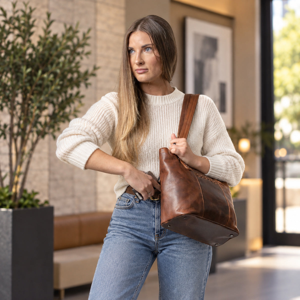 A woman wearing a cream ribbed knit sweater and blue jeans carries a large brown leather shoulder bag in a modern indoor setting. Women Lady Conceal - YKK Locking & Universal Holster - Designer Luxury Leather Carry Handbag - carry Handbag for gun carry - Unique Tote gun Handbag