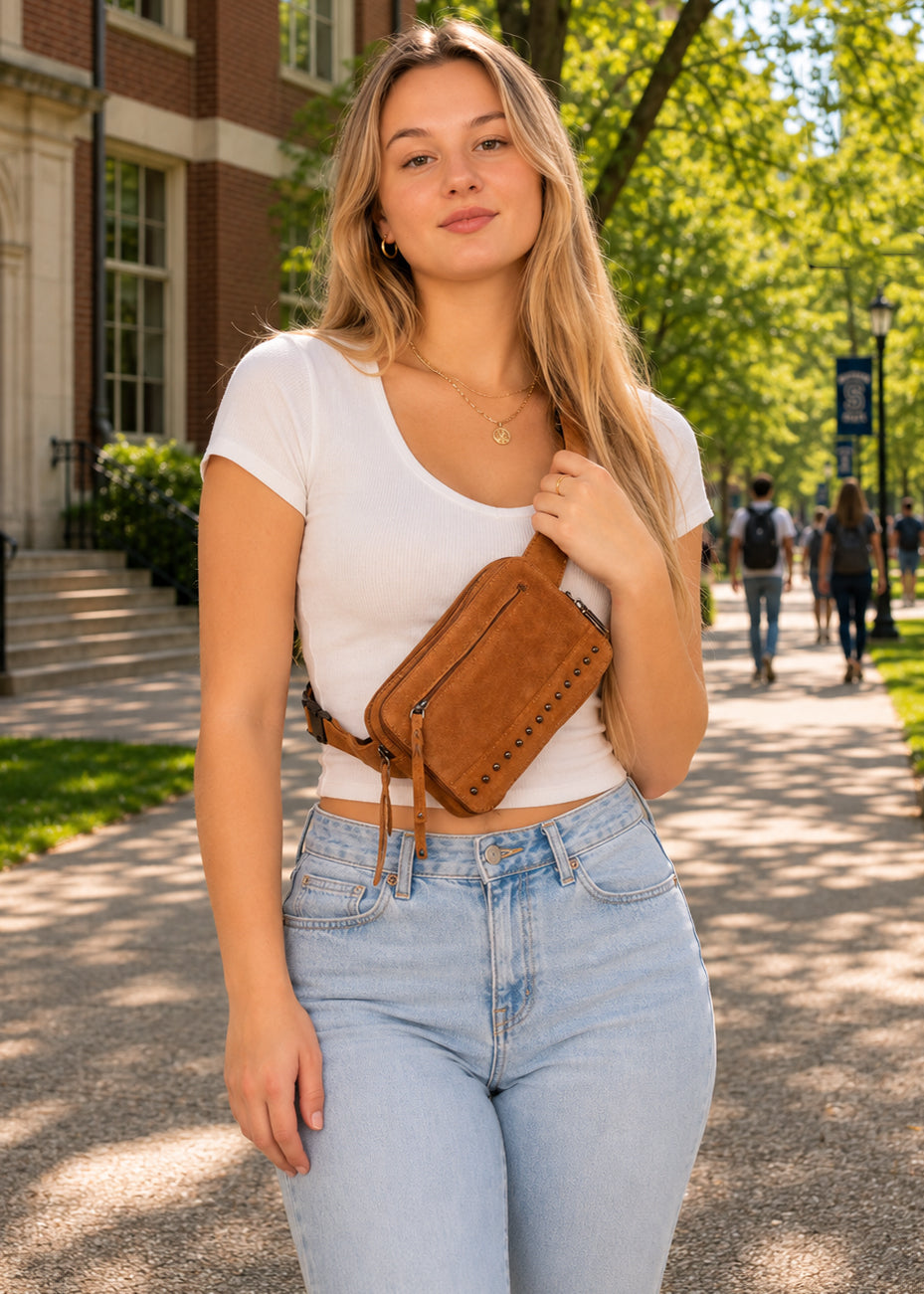A young woman stands on a sunlit campus walkway wearing a white cropped T-shirt, light blue jeans, and a brown suede crossbody bag with a zipper and tassel detail.